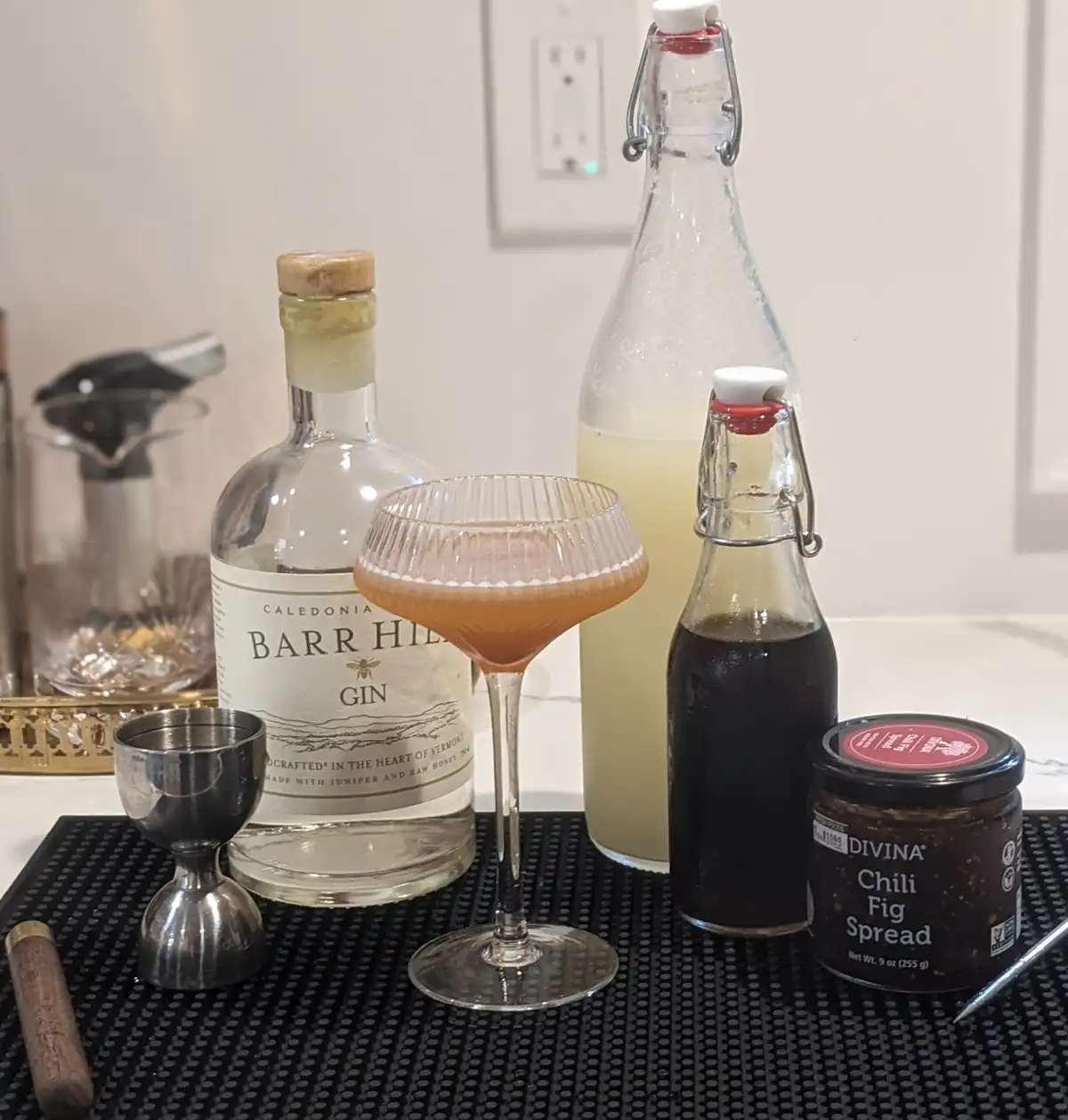 A cocktail in a coupe glass sits on a black bar mat surrounded by a bottle of Barr Hill Gin, a jar of Divina Chili Fig…