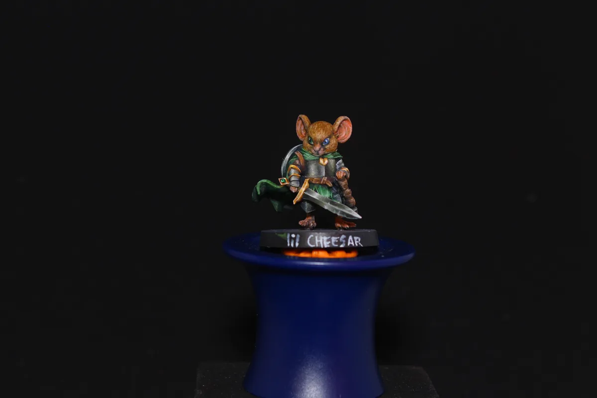 A painted miniature of a mouse warrior in armor and a green cape, holding a sword and shield, stands atop a blue magician's…