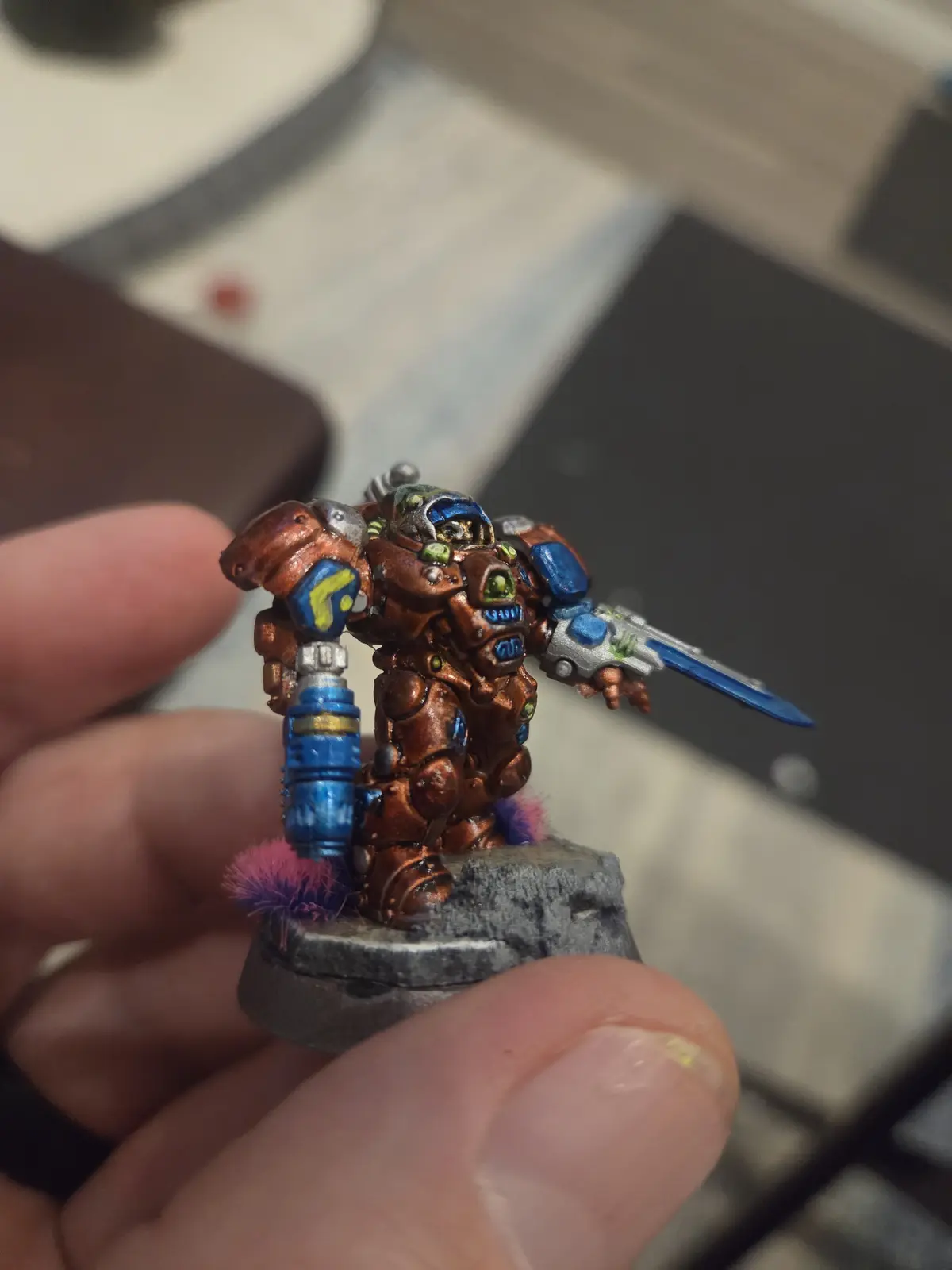 A hand holds a painted miniature of a heavily armored, biomechanical warrior with coppery-brown armor, blue and yellow…