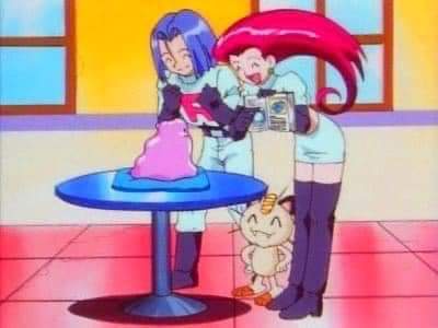 Two anime characters stand beside a round blue table with a pink cake on it. The character on the right, with long red hair…