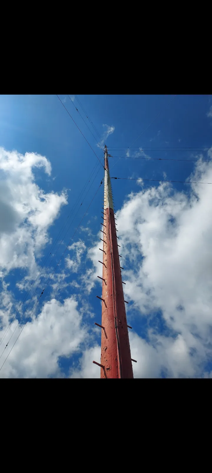 A low-angle photo shows a tall, red-painted utility pole stretching upward against a blue sky with white clouds. Several…