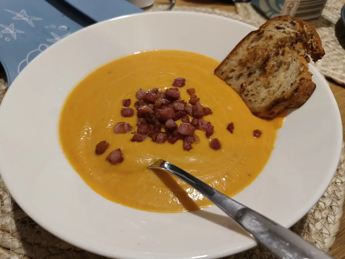 A white bowl contains creamy orange soup topped with crispy bacon bits and a slice of toasted bread on the side. A spoon…