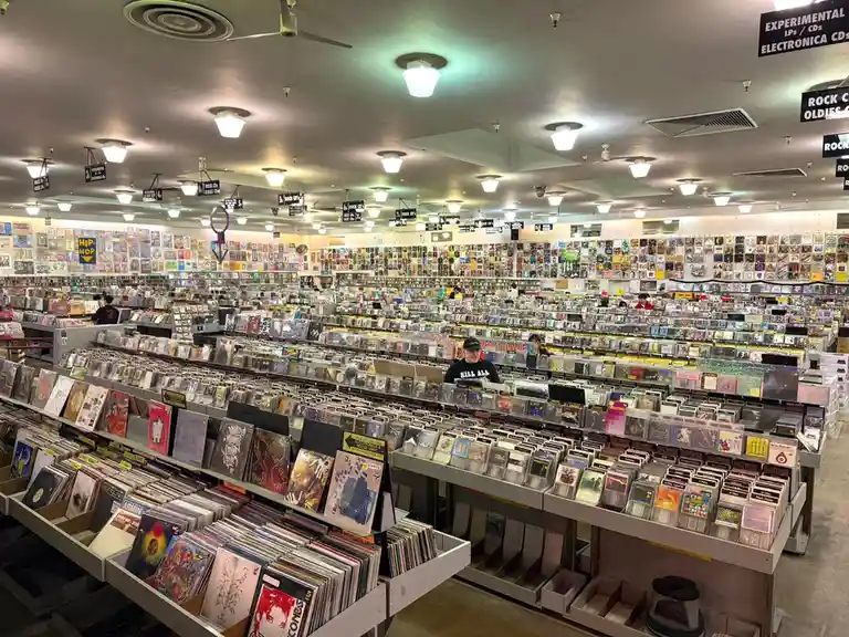 An expansive record store filled with rows upon rows of vinyl records and CDs, with overhead signs categorizing genres like…