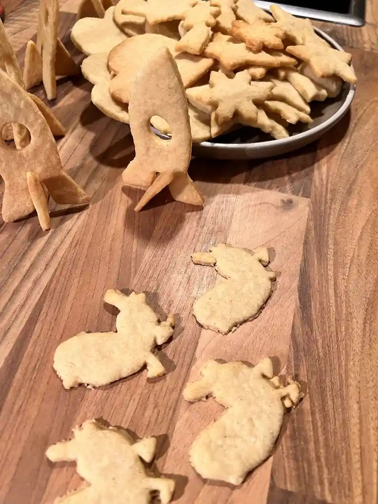 A wooden table displays homemade cookies: several rocket-shaped cookies stand upright in the foreground, while a plate piled…