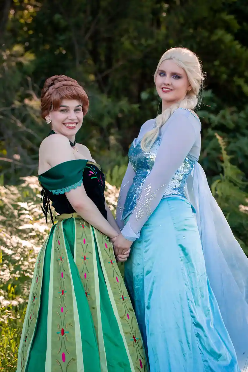 Two women dressed as Disney's Anna and Elsa from Frozen stand side by side outdoors, holding hands and smiling at the…
