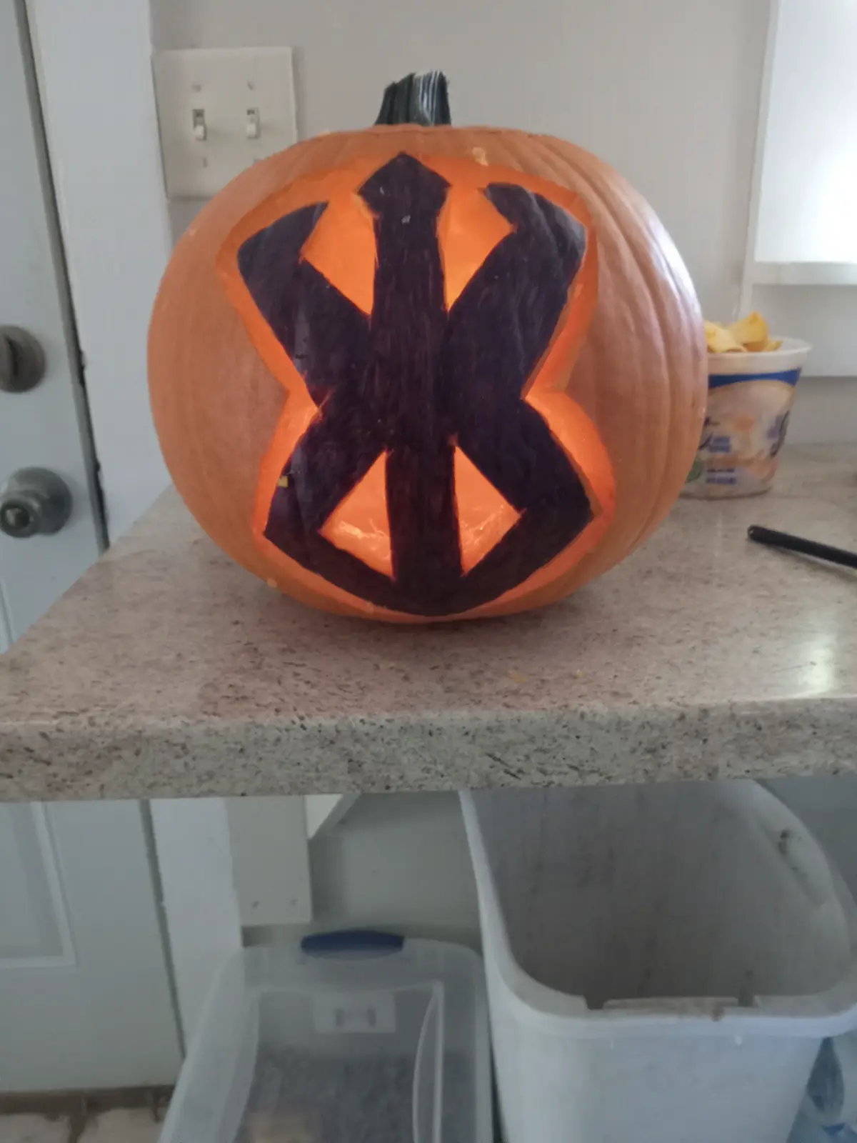 A carved pumpkin sits on a kitchen counter, illuminated from within, with a large black peace symbol painted over its carved…