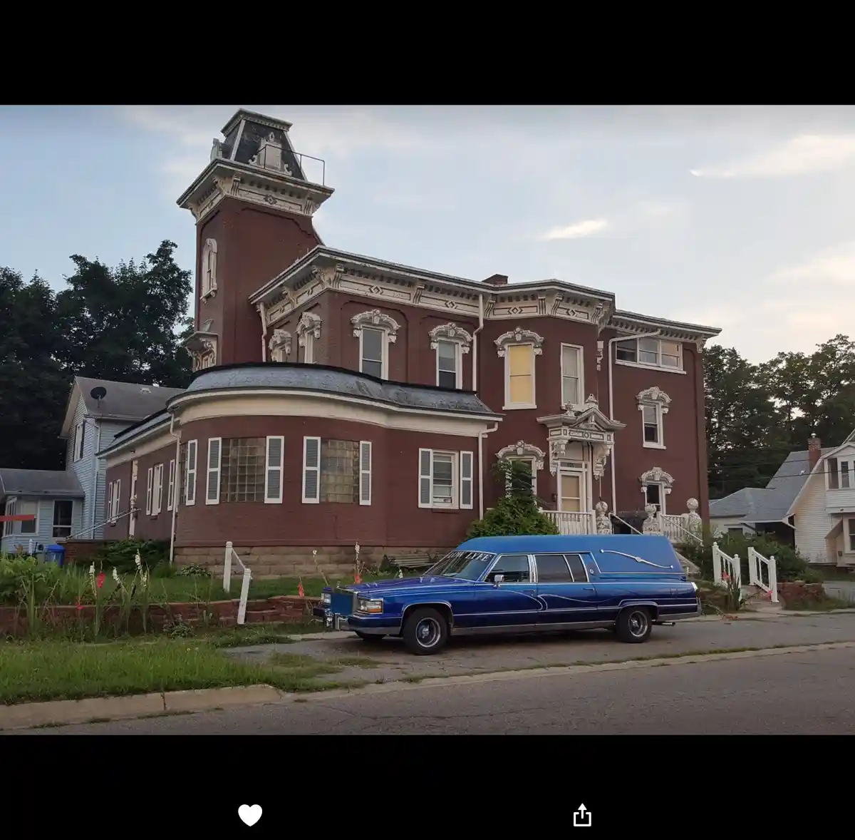 A blue hearse is parked in front of a large, ornate, red brick Victorian-style house with white trim and a tower. The text…