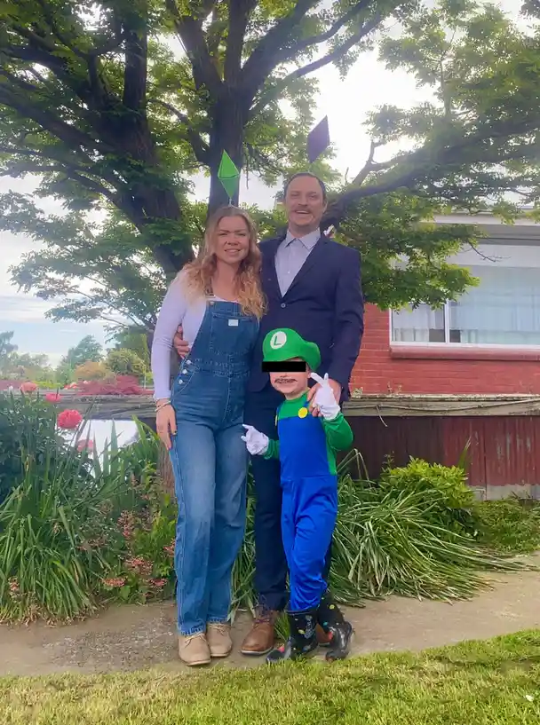 A family photo shows a woman in denim overalls, a man in a suit, and a child dressed as Luigi from Mario games, standing…
