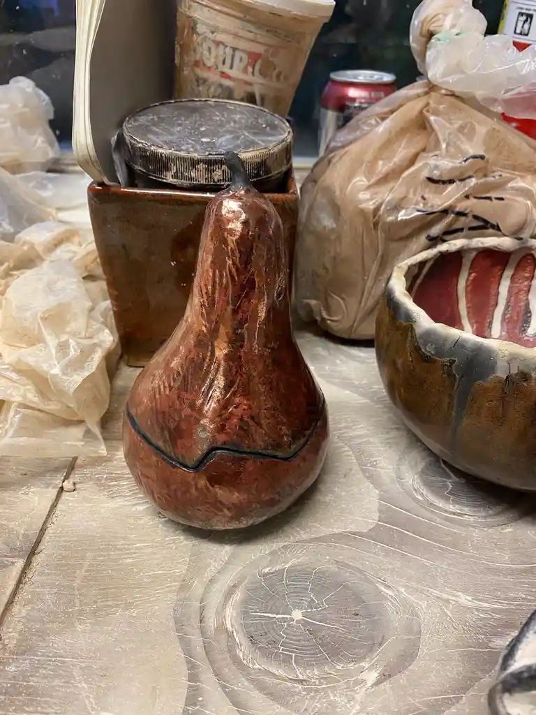 A ceramic gourd-shaped vase with a prominent black crack across its midsection sits on a worn wooden table. Surrounding it…