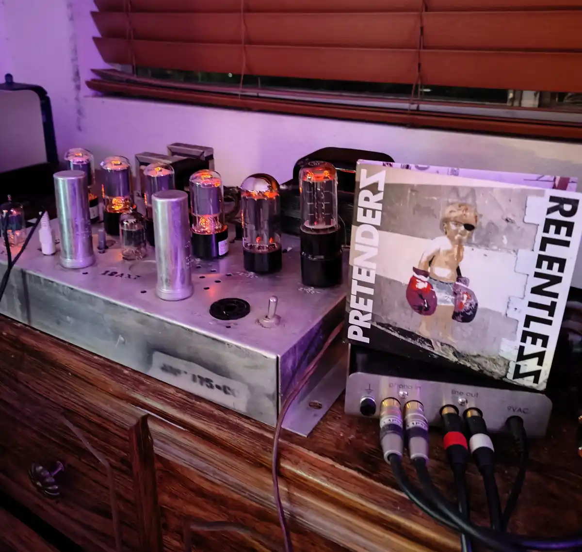 A close-up photo shows a vintage tube amplifier with glowing vacuum tubes on a wooden surface, next to a vinyl record sleeve…