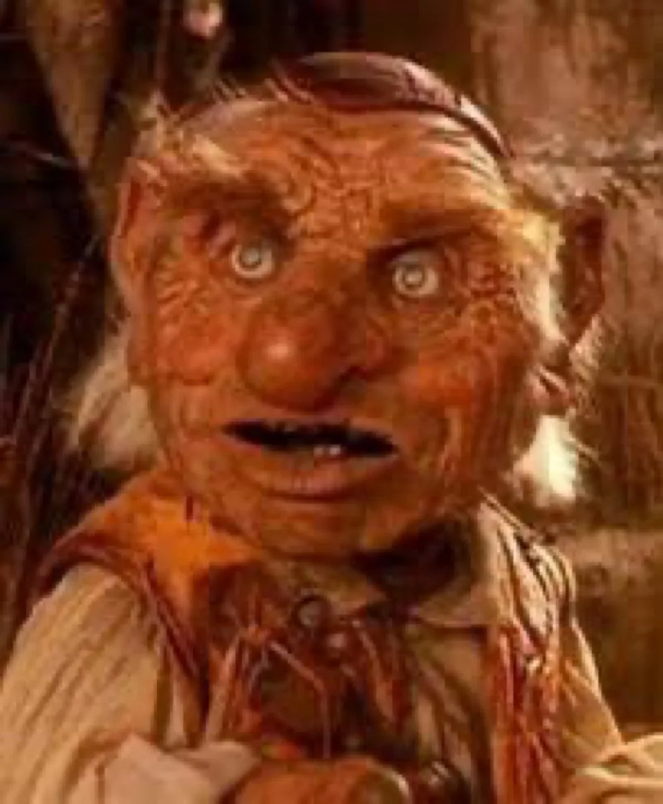 A close-up of a CGI-rendered, orange-skinned, wrinkled creature with wide, startled eyes and an open mouth, wearing a…