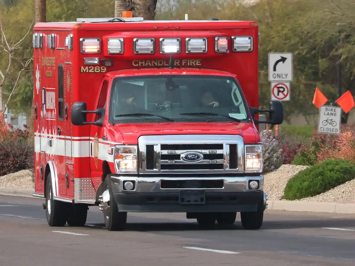 A red Chandler Fire ambulance, marked M289, is driving on a street with its emergency lights flashing. Visible signs include…
