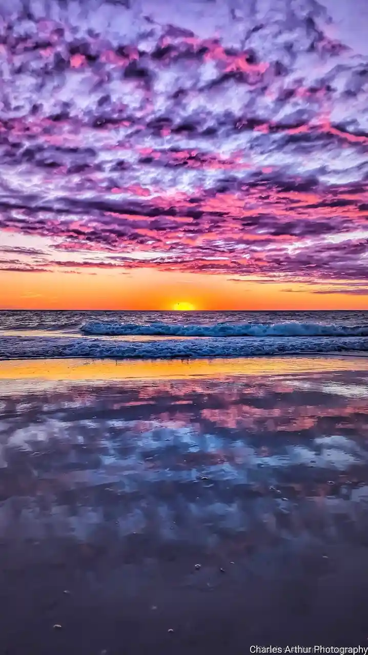 A stunning photograph of a beach sunset with vibrant purple and pink clouds reflecting in wet sand, the sun setting on the…