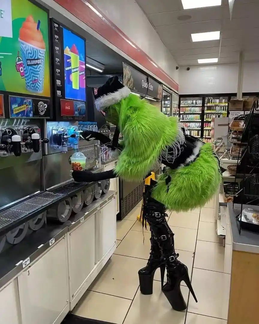 A person in a full-body green Grinch costume, complete with a Santa hat, is standing on extremely high black platform heels…