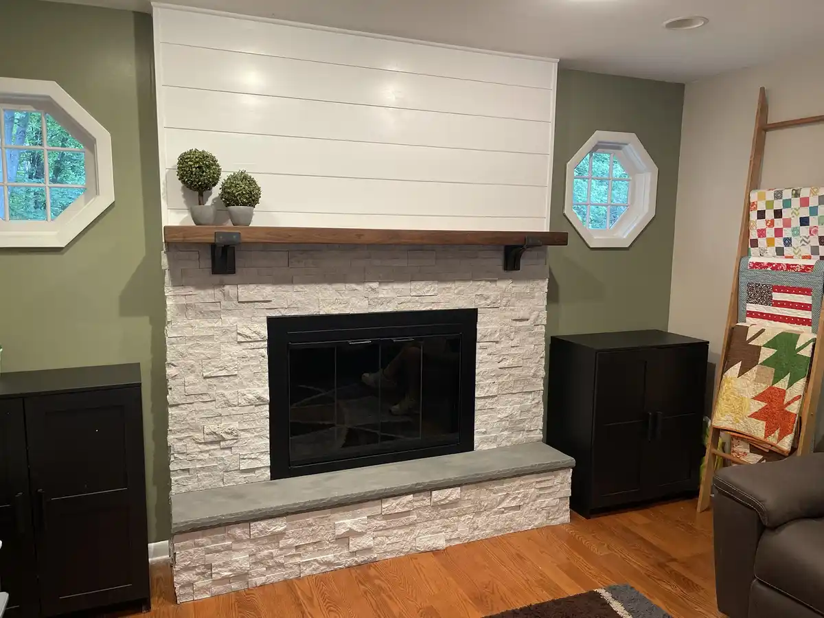 A modern fireplace with a white shiplap wall above, a wooden mantel holding two small potted plants, and a stone surround…