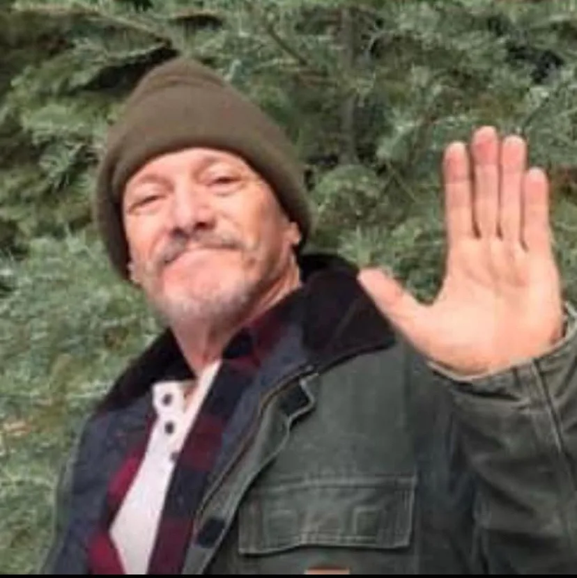 A middle-aged man with a mustache and beard, wearing a brown beanie, a plaid shirt under a dark jacket, stands outdoors in…