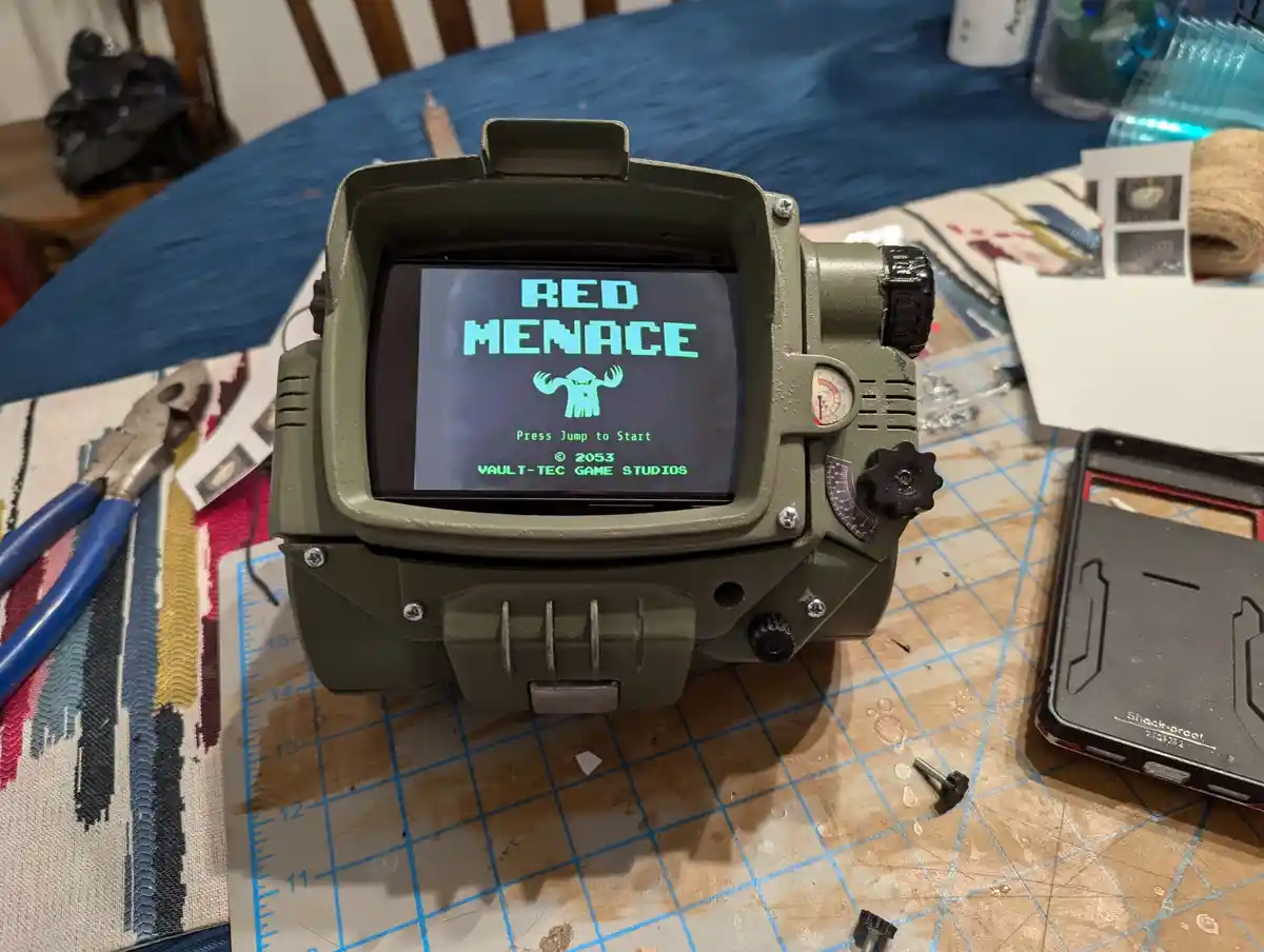 A green, rugged handheld device resembling a Fallout Pip-Boy sits on a cluttered workbench with tools and a cutting mat. Its…
