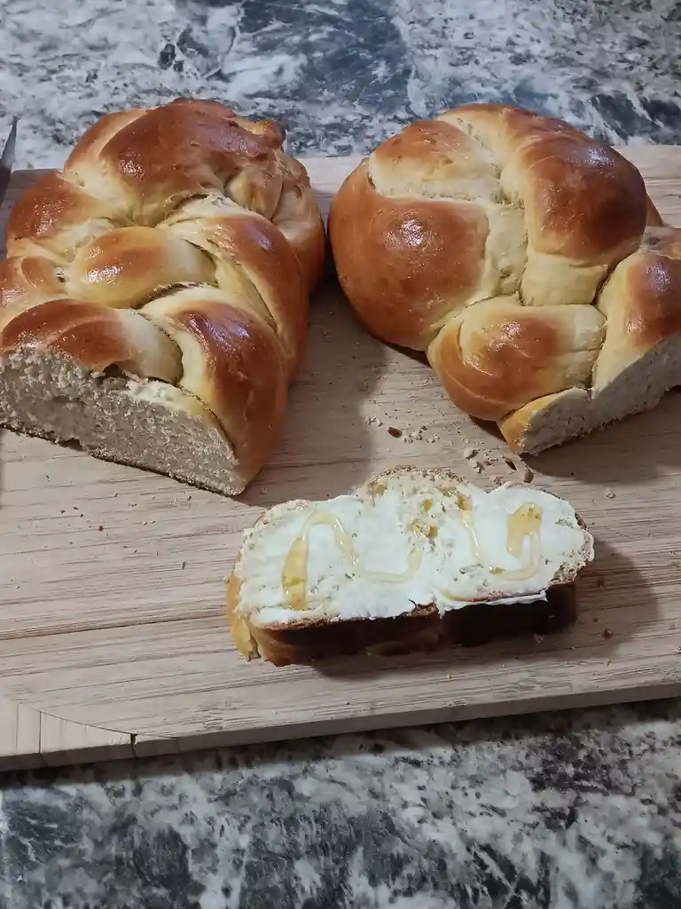 Two braided loaves of golden-brown bread sit on a wooden cutting board, one sliced open to reveal soft crumb. In the…