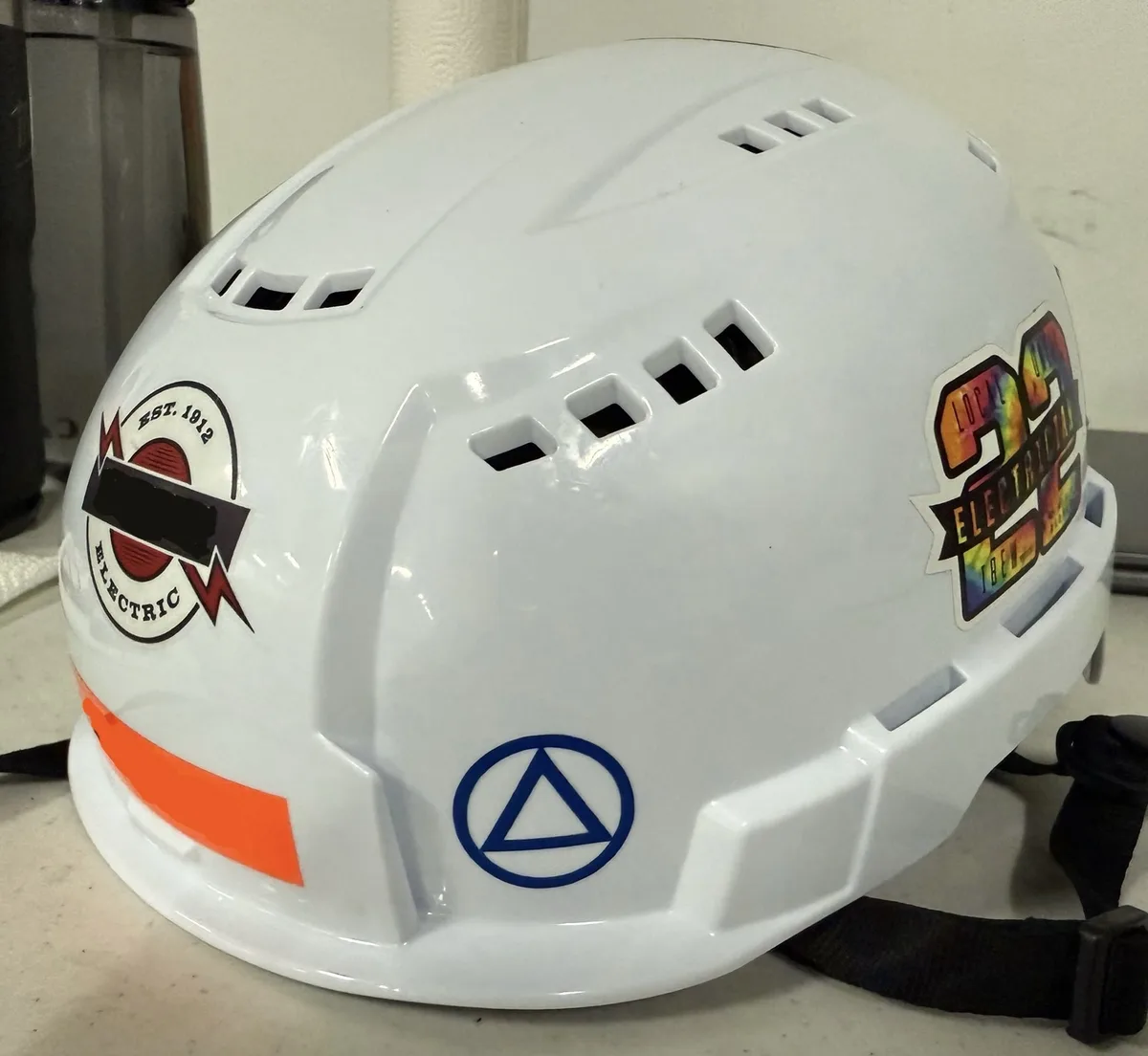 A white hard hat with ventilation slots and a black strap rests on a surface. It features a red and black 'EST. 1912…