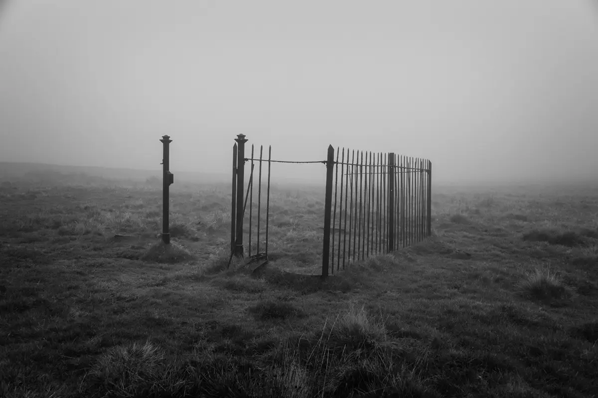 A black-and-white photo shows a rusty, broken metal gate in a foggy, grassy field. The gate is partially open and appears…