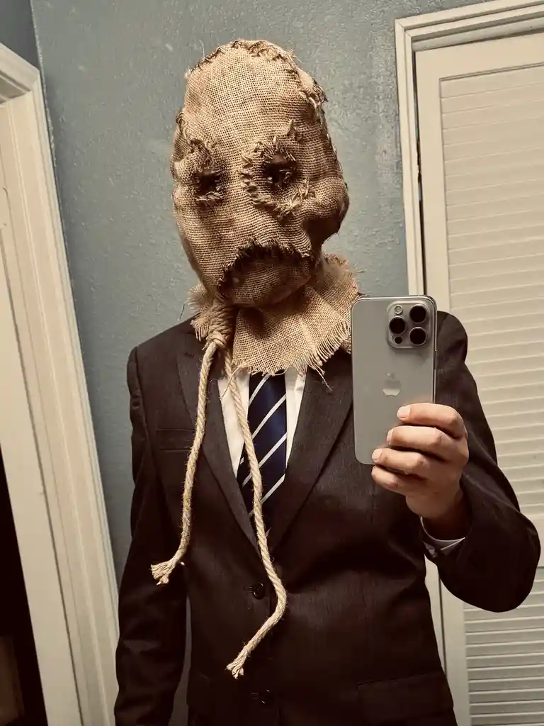 A person wearing a dark suit, white shirt, and striped tie takes a mirror selfie with a silver smartphone. Their face is…