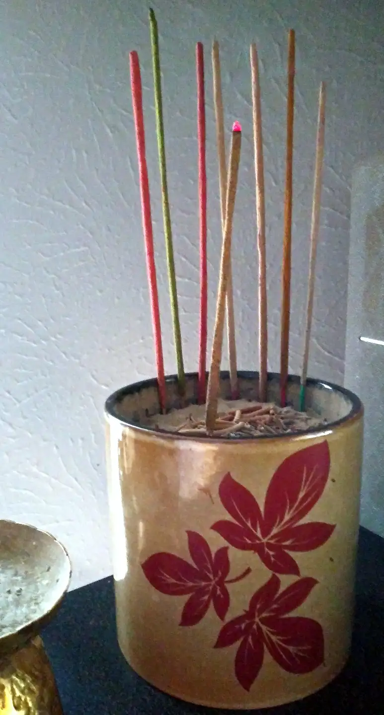 A ceramic incense holder with red leaf patterns contains multiple incense sticks; one stick is lit with a glowing tip while…