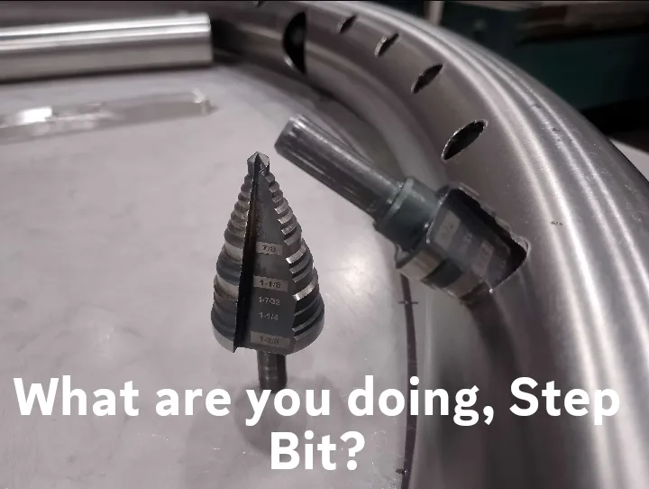 A close-up photo shows a metal step drill bit standing upright on a workbench next to a curved metal pipe with holes. The…