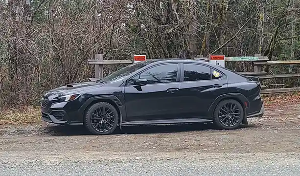 A dark gray Subaru WRX sedan is parked on a gravel surface in front of a wooden fence and bare trees. Two orange and white…