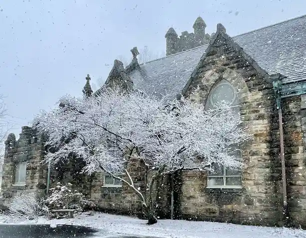 A snow-covered stone church with Gothic architectural details, including pointed arches and crosses, is shown during a…