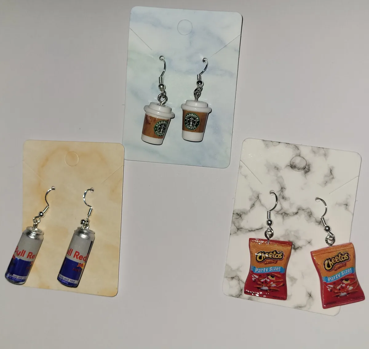 Three pairs of novelty earrings are displayed on separate cards: Red Bull energy drink cans, Starbucks coffee cups, and…