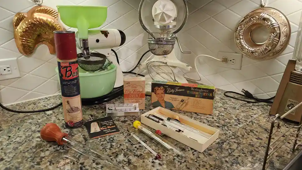 A kitchen countertop displays a collection of vintage cooking tools and thermometers, including a green stand mixer, a can…