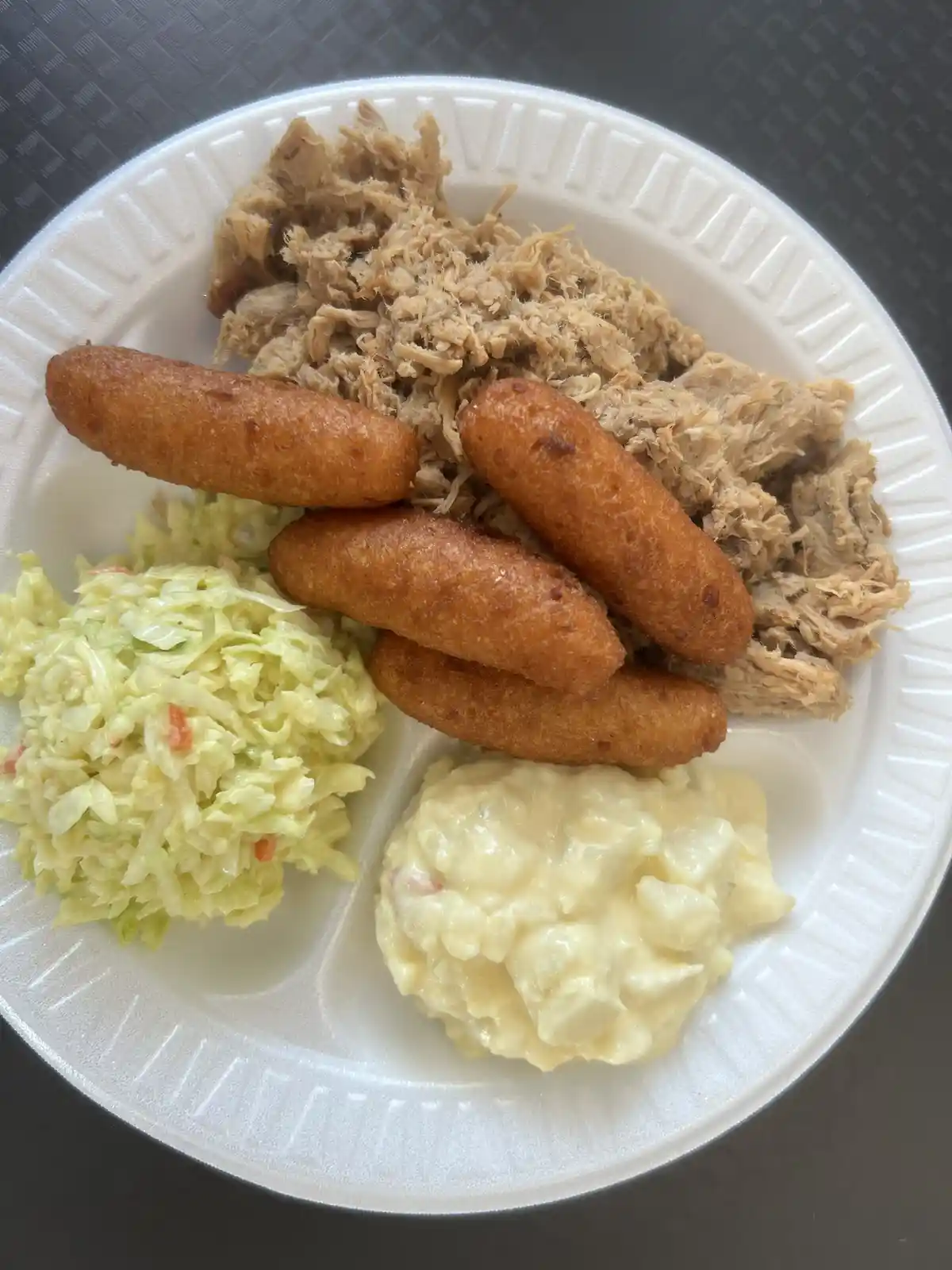 A white styrofoam plate holds a meal of shredded meat, four golden-brown corn dogs, coleslaw, and potato salad. The overlaid…