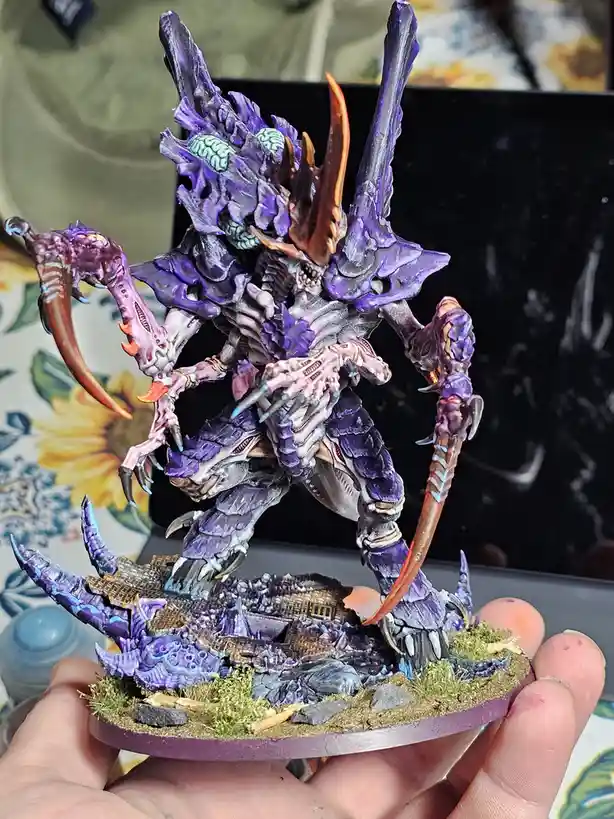 A person holds a highly detailed, purple-painted miniature of a monstrous, multi-limbed alien creature with glowing green…