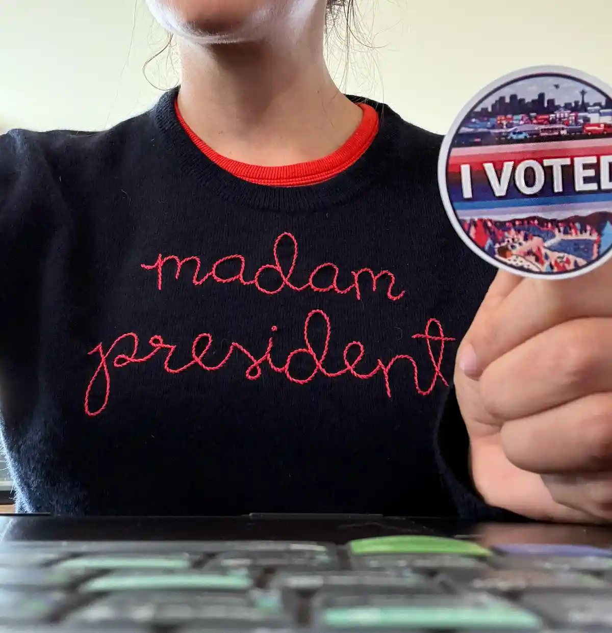A person wearing a black sweater with 'madam president' embroidered in red cursive holds up an 'I VOTED' sticker, implying…
