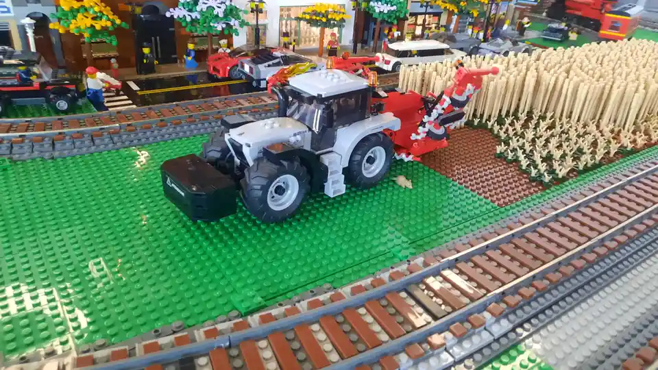 A detailed LEGO diorama features a white and red tractor with a harvesting attachment on a green baseplate, positioned…