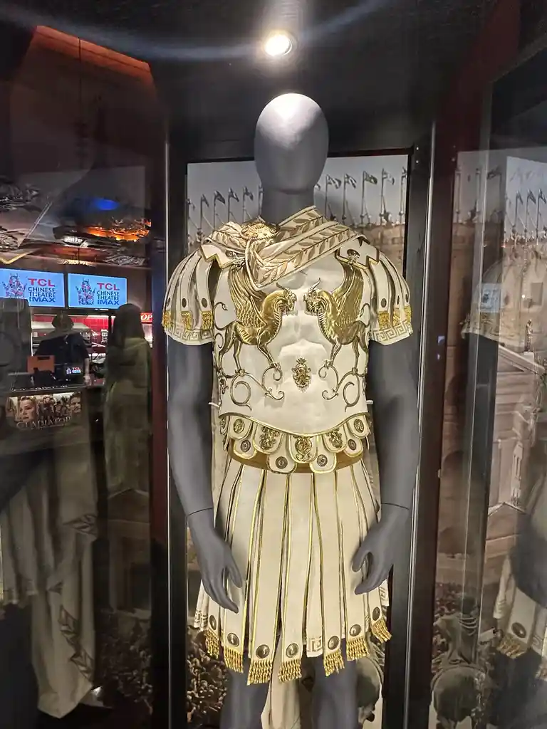 A mannequin in a glass display case wears an ornate, gold-embellished Roman-style costume with griffin motifs, resembling…
