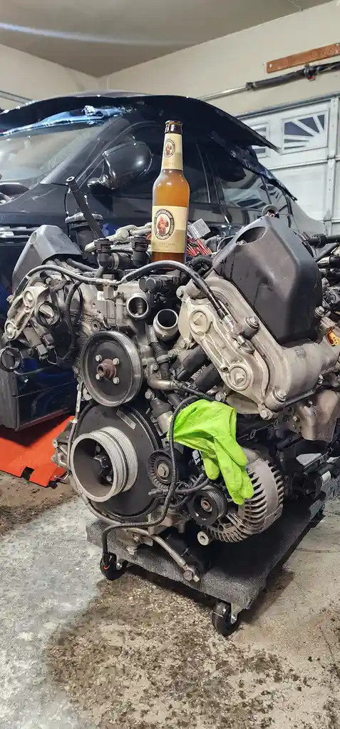 A disassembled car engine sits on a wheeled cart in a garage, with a bottle of Franziskaner Weissbier beer balanced on top…