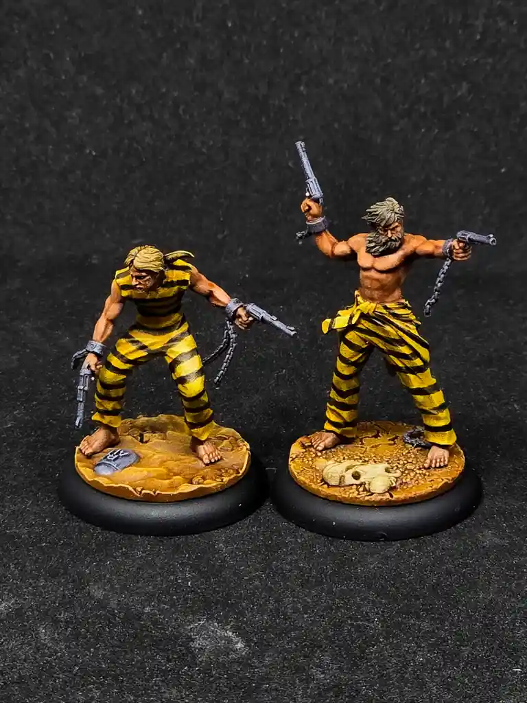 Two painted miniature figures in yellow and black striped prison uniforms, each holding a pistol and a chain, stand on…