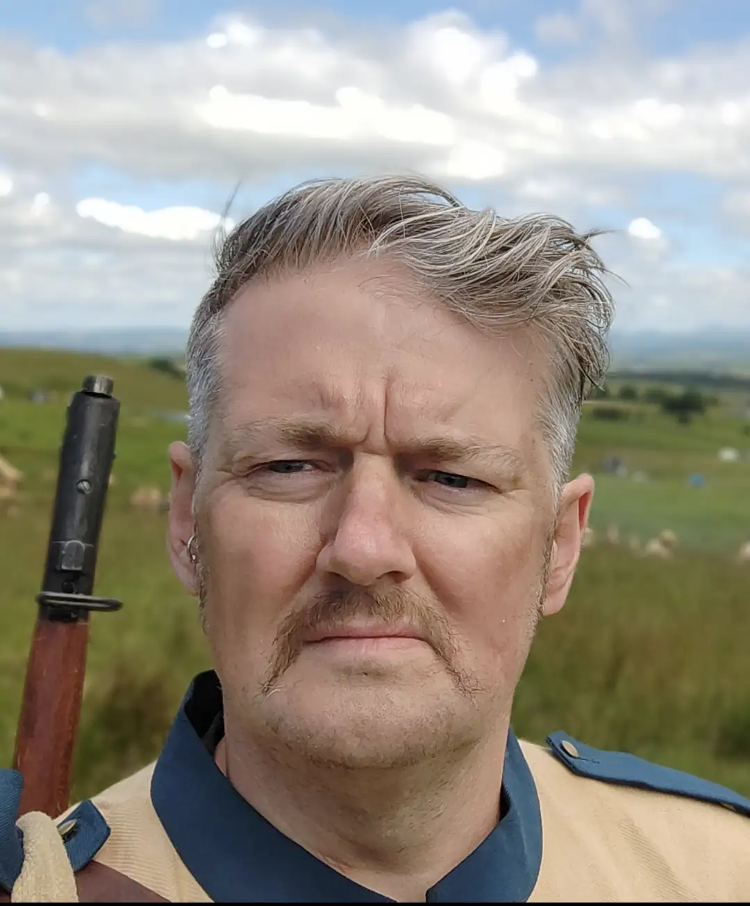 A man in a beige and blue historical military uniform with a rifle slung over his shoulder stands in a grassy field under a…
