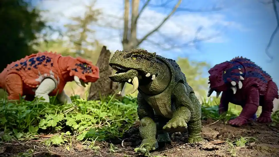 Three plastic dinosaur toys are posed in a grassy outdoor setting, with a green one in the center looking forward, flanked…