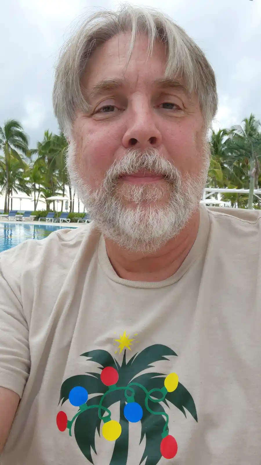 A man with a white beard and gray hair takes a selfie in front of a pool with palm trees in the background. He wears a beige…
