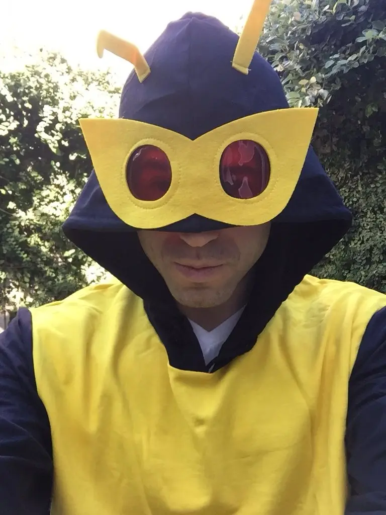 A person wearing a homemade costume resembling a cartoon bee, featuring a dark blue hood with yellow antennae, a large…