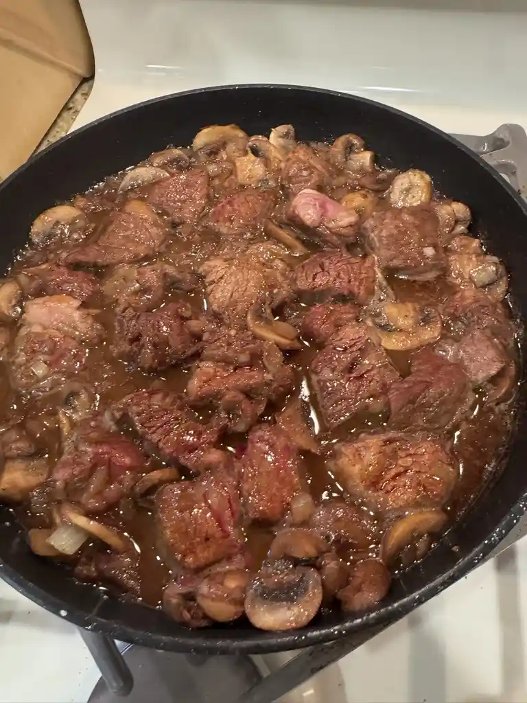 A close-up photo shows a black skillet on a stovetop filled with chunks of raw-looking meat, sliced mushrooms, and onions in…