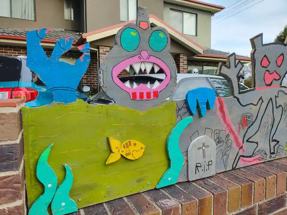 A handmade Halloween decoration on a brick wall features a large grey monster with green eyes and pink mouth, blue clawed…