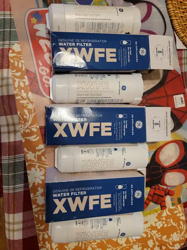 A stack of six GE refrigerator water filter boxes and filters, labeled 'XWFE', arranged vertically on a colorful patterned…