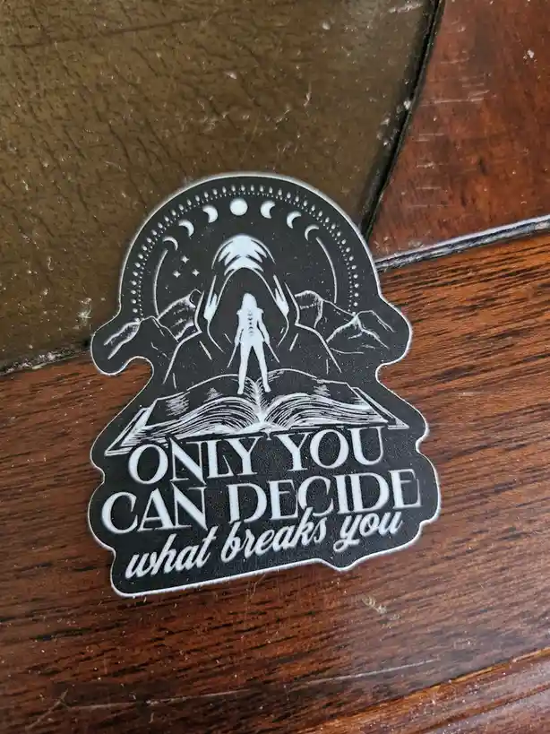A black and white sticker on a wooden surface features a silhouette of a person standing atop an open book, framed by…