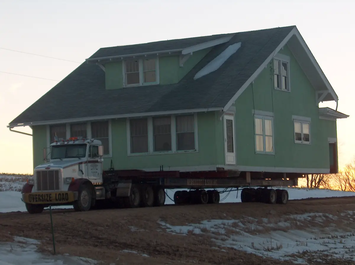A large white semi-truck with a yellow 'OVERSIZE LOAD' sign is hauling a two-story green house on a flatbed trailer across a…