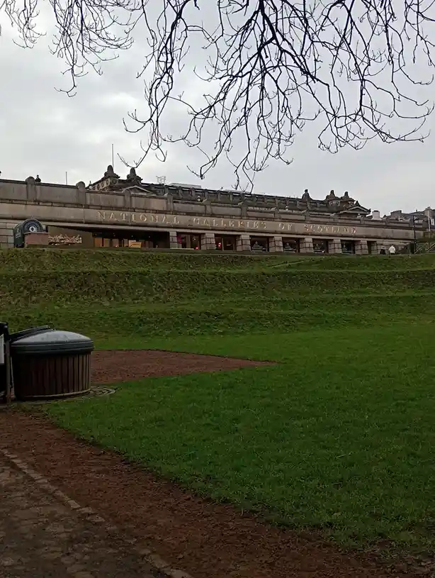 A photo of the National Galleries of Scotland building, viewed from a grassy slope with bare tree branches framing the top…