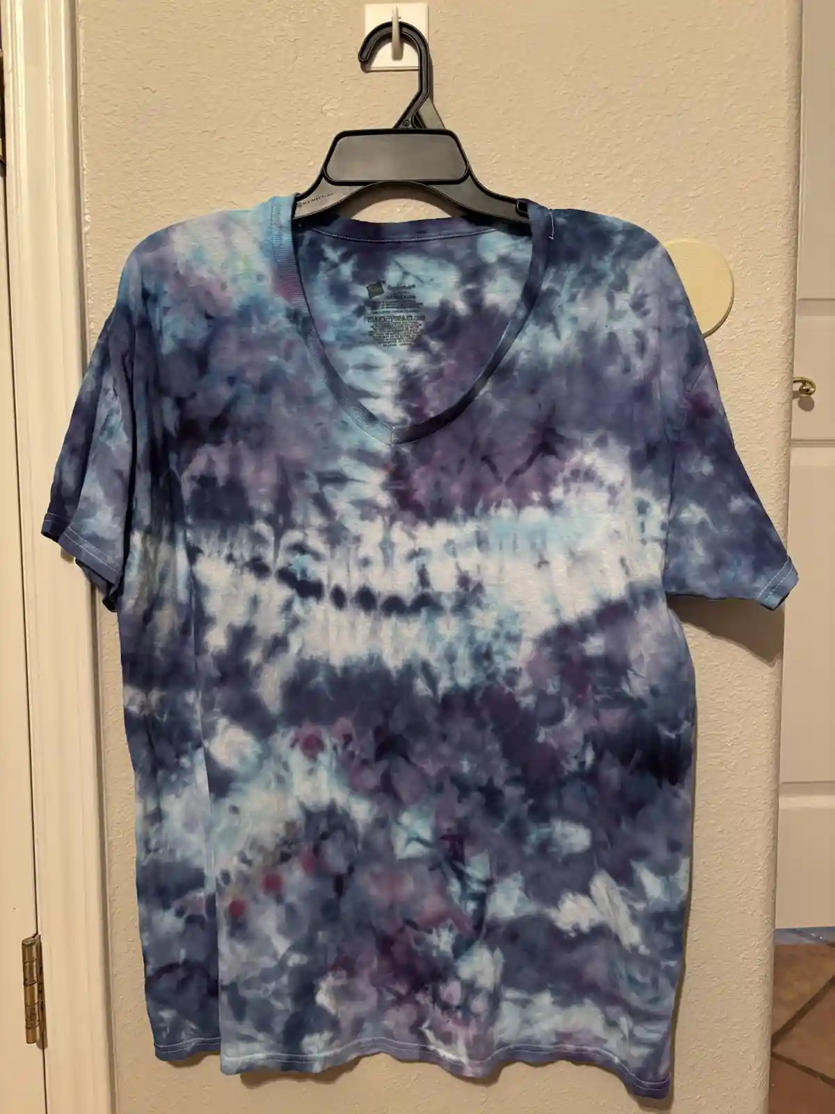 A v-neck tie-dye t-shirt with swirling blue, purple, and white patterns hangs on a black plastic hanger against a beige…