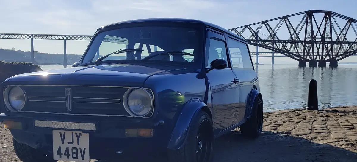 A dark blue classic Mini car is parked on a cobblestone waterfront, with a large steel truss bridge spanning the water in…